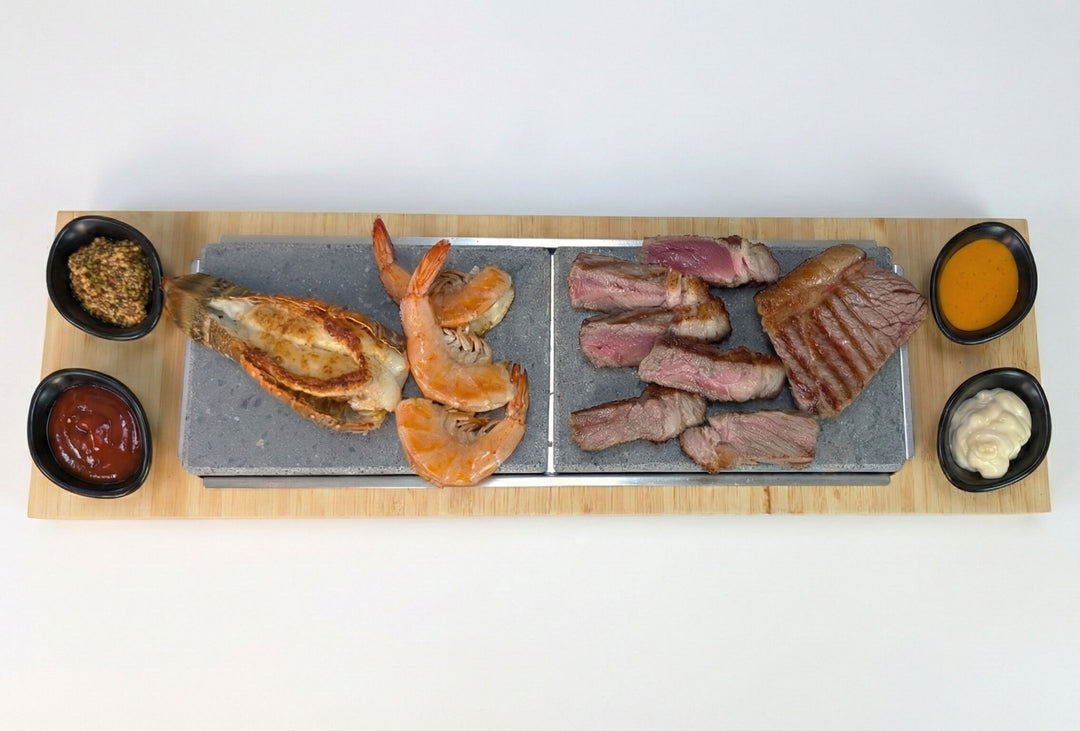 The Black Rock Grill Sharing Lava Sizzler Finishing Warming Platter features a rectangular wooden tray and natural lava stone slabs, served with grilled lobster, shrimp, sliced steak, and four bowls of various sauces and dips.