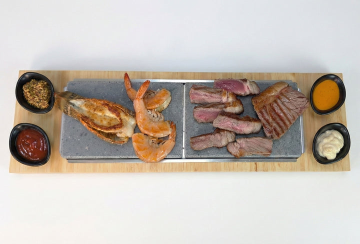 The Black Rock Grill Sharing Lava Sizzler Finishing Warming Platter features a rectangular wooden tray and natural lava stone slabs, served with grilled lobster, shrimp, sliced steak, and four bowls of various sauces and dips.