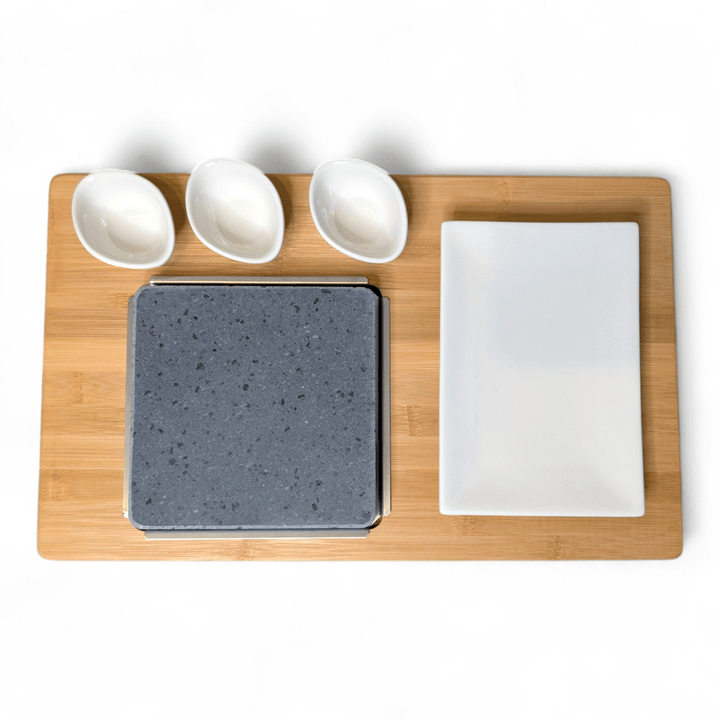 A bamboo serving board set on a white background, featuring a square lava cooking stone, a rectangular white side plate and three small white dipping pots.
