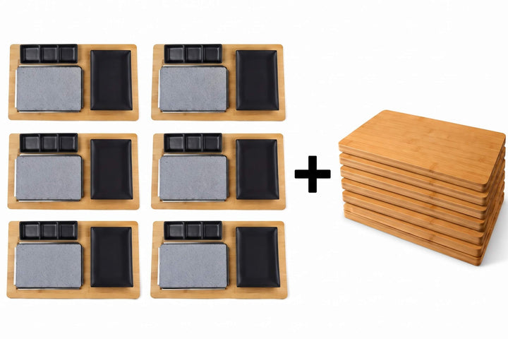 Six sets of Black Rock Grill Black Steak Stone Cooking Sets, each featuring a rectangular wooden tray, stone slab, black dishware, and a FREE £19.99 Bamboo Board, are arranged beside a stack of six empty wooden trays on a white background. Perfect for hot stone cooking at home or in restaurants, these versatile sets deliver the authentic Black Rock Grill experience to any meal.