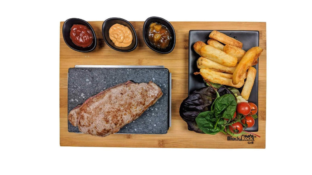 The Black Rock Grill Case of 6 Steak Stone Lava Cooking Stone Rectangle Rocks ROK-06 presents steak sizzling on the hot rock, served with fries, fresh tomato salad, and three sauces arranged neatly on a wooden board.