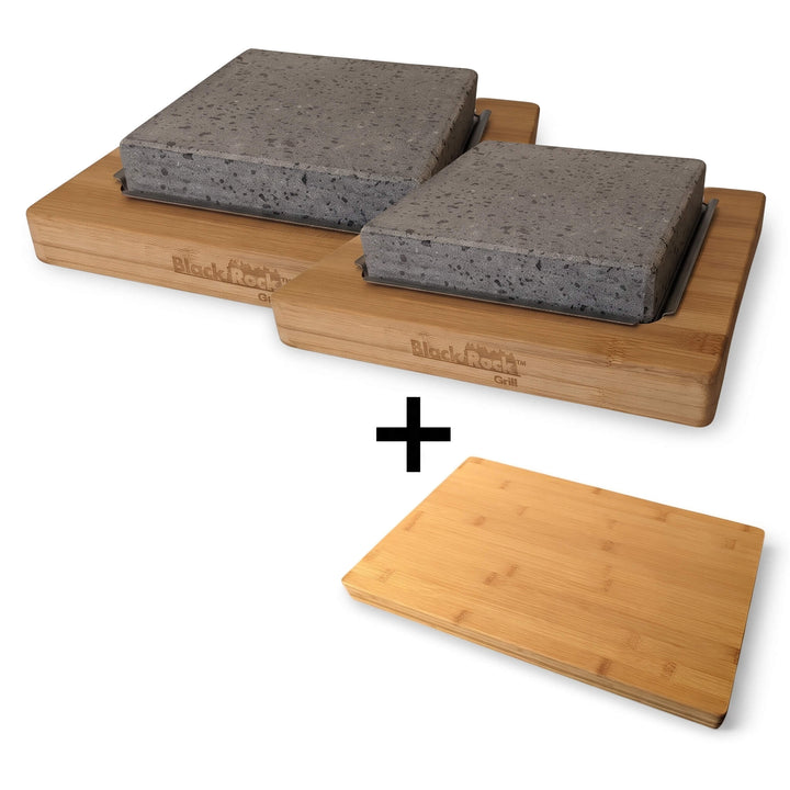 Side by side, two rectangular bamboo boards from Black Rock Grill each hold a large gray lava stone slab, forming the NEW Black Rock Grill Lava Stone Steak Multi Pack. Beneath them, a single complimentary bamboo board (worth £19.99) completes this elegant hot stone cooking set, separated by a black plus sign.