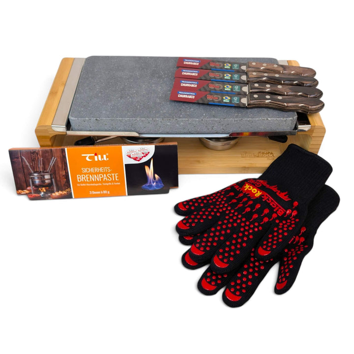 A bamboo-framed tabletop hot lava stone cooking set stove with two stainless steel gel burners to keep the stone hot for longer, with a pack of till brenpaste chaffing gel fuel in the foreground, as well a pair of heat resistant gloves,four wrapped tramontina steak knives are rested on the lava stone, photographed on a clean white background