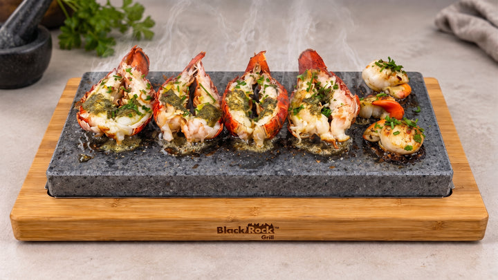 Black Rod Grills hot stone platter with lobster tails and scallops cooking over visible steam, set on a bamboo board against a light stone countertop.