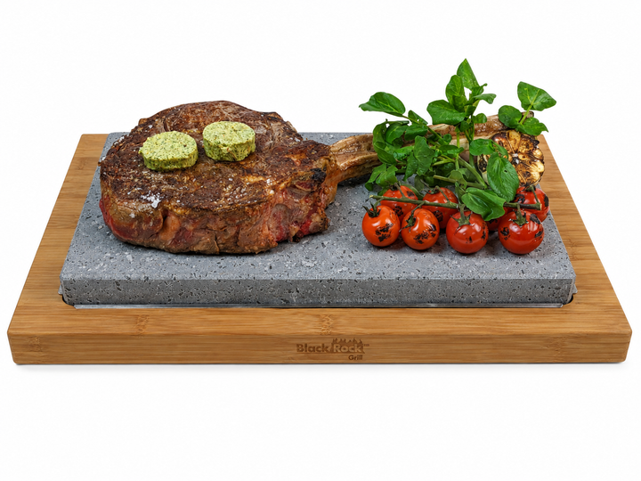 A rectangular bamboo serving board with a lava cooking stone insert, cooking a large tomahawk steak, watercress and a cluster of red cherry tomatoes on a white background.