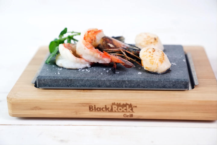 A Black Rock Grill branded warming stone cooking set holding cooked shrimp, scallops, and a sprig of green herb on a white background.