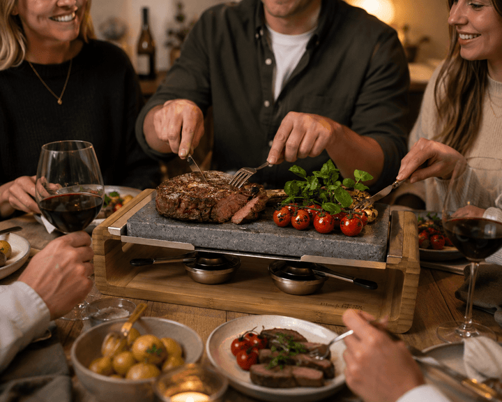 A group of friends gathered around a table, sharing a cooked steak on a stone plate with cherry tomatoes and herbs, set in a warm, cozy dining environment.