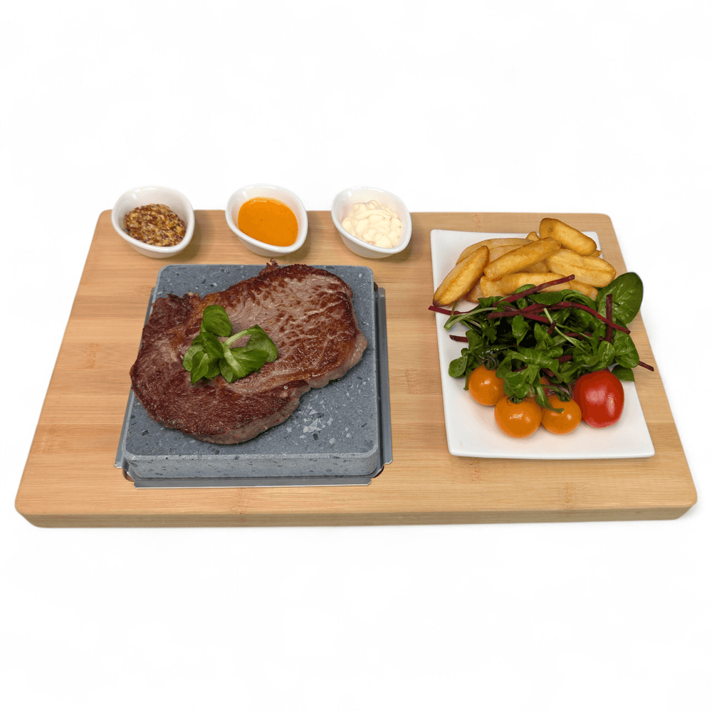 A wooden serving board with a Lava cooking stone holding a sizzling steak topped with green garnish, alongside a white rectangle side plate with fries and salad and three small dipping pots of seasoning, sauce, and cheese on a white background.