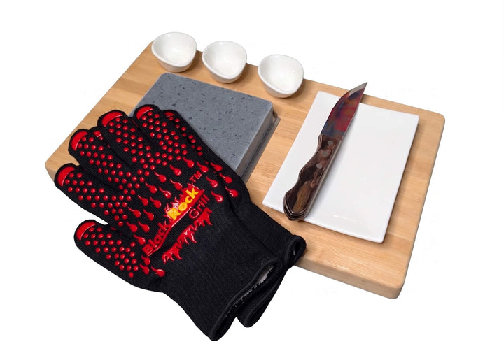 A bamboo serving board set on a white background, featuring a square lava cooking stone with a pair of black rock grill heat resistant gloves placed over one corner, a rectangular white side plate with a steak knife on it and three small white dipping pots.