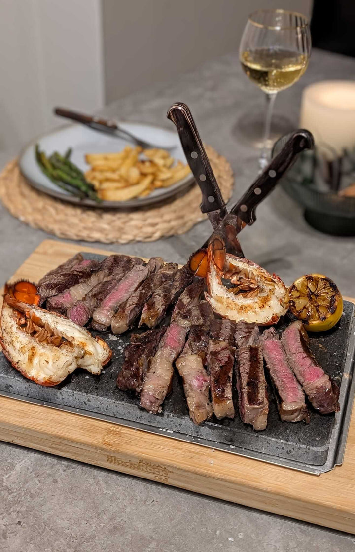 A platter of sliced medium-rare steak on a Black Rock Grill Large Lava Sizzler Finishing Warming Platter with two steak knives, served with grilled lobster tails, charred lemon halves, fries, asparagus, and a glass of white wine.