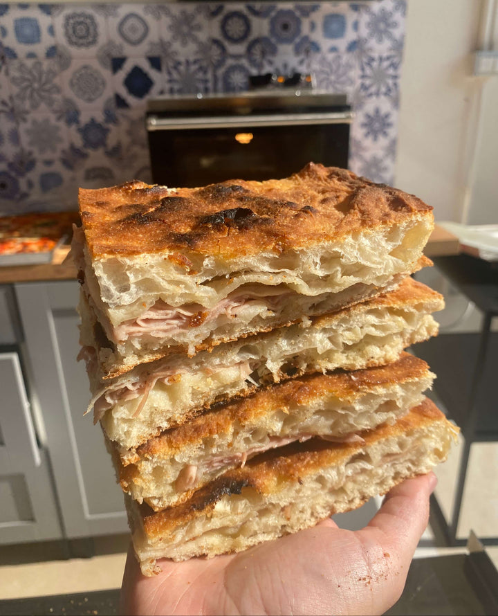 A hand holds three thick sandwiches stacked high with golden focaccia bread, made in Italy—perfect for baking in the Macte Nettuno Electric Pizza Oven by Macte. A kitchen with a blue tile backsplash is seen in the background.