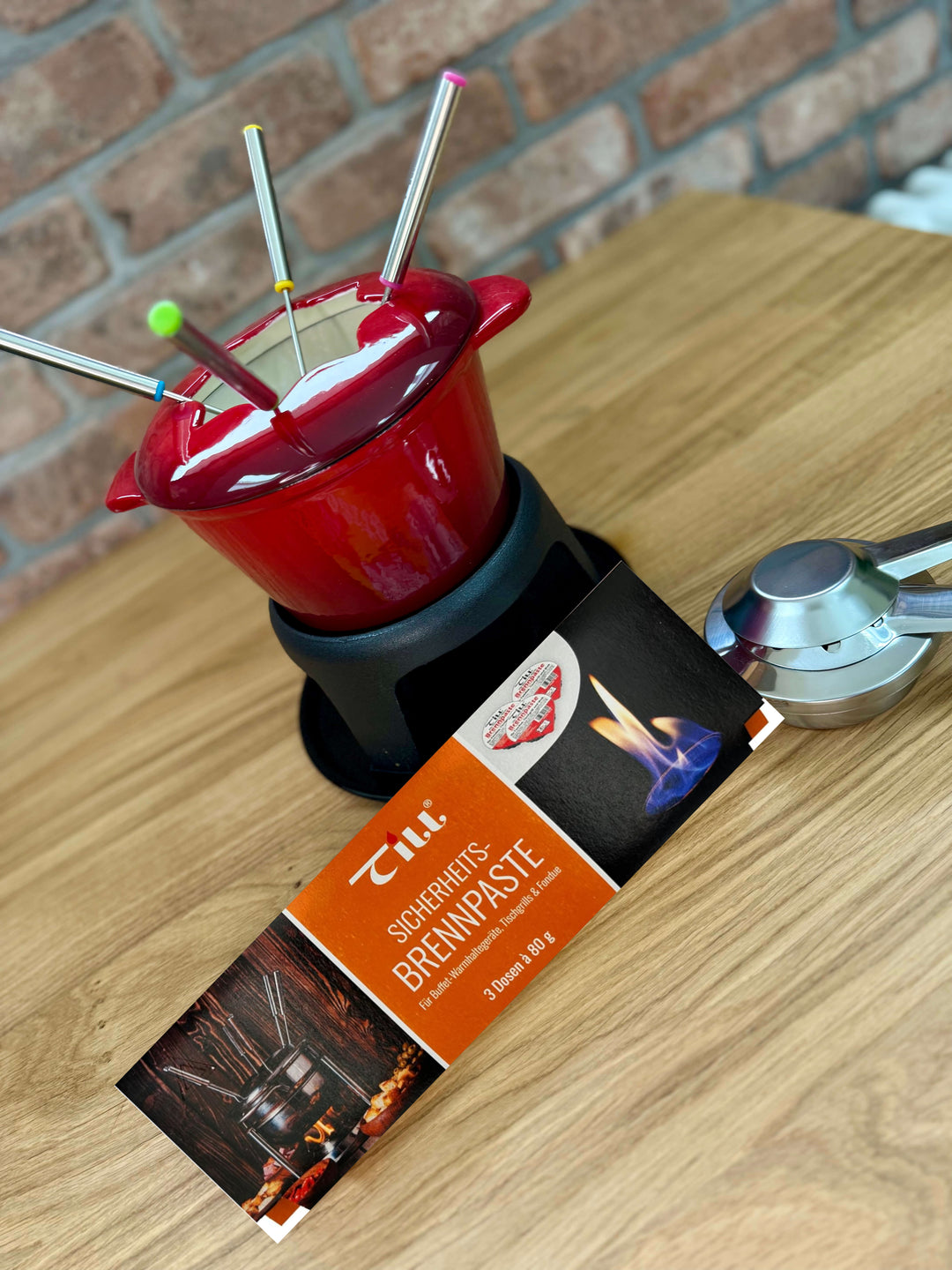 A red fondue pot with four colorful forks sits on a wooden table. In front are a Till Brennpaste Fondue Fuel Gel box and metal container by Black Rock Grill, with a brick wall background for cozy fondue vibes.