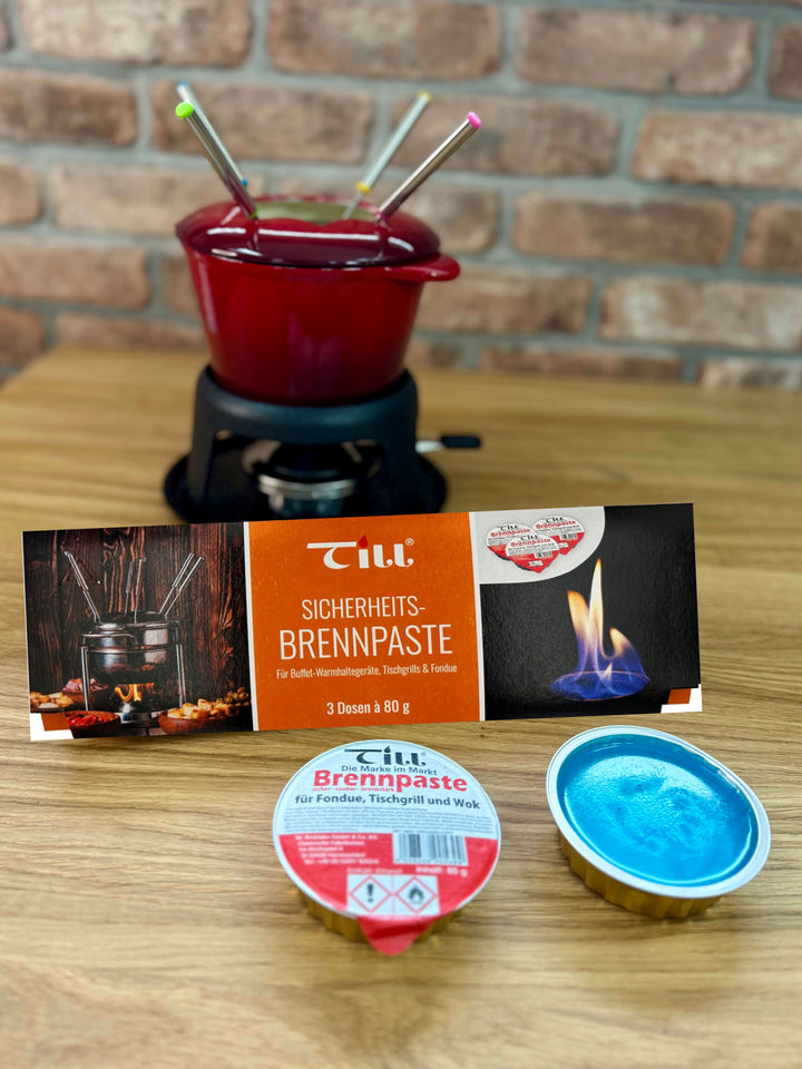 A red fondue pot with four forks on a black stand is in the background. In front are a package of Till Brennpaste Fondue Fuel Gel and two blue tins of Black Rock Grill fondue gel, all placed on a wooden surface.