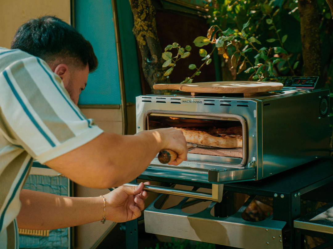 A cook removing a freshly baked pizza from the Macte Voyager Twin Advanced oven in an outdoor garden setting.