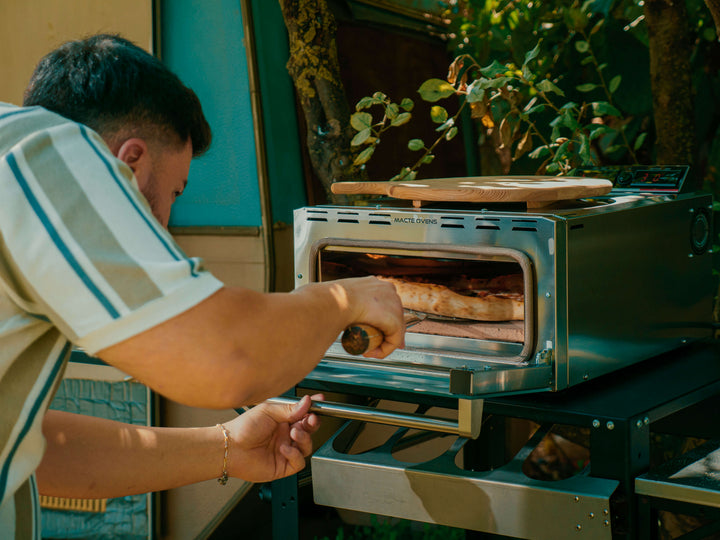 A cook removing a freshly baked pizza from the Macte Voyager Twin Advanced oven in an outdoor garden setting.