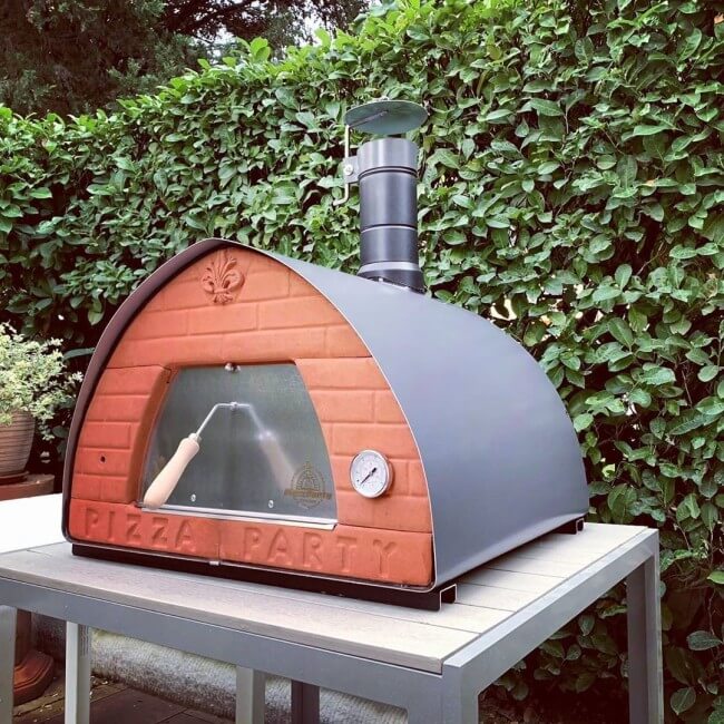 Stone 70X70 Pizza Party outdoor pizza oven with brick design and stone color finish on a table against a green hedge background.