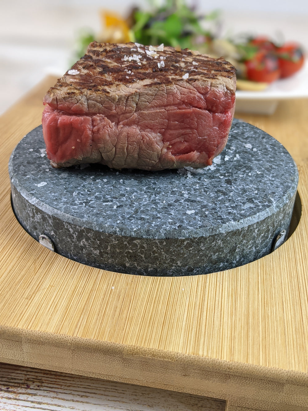 A thick slice of rare steak with a seared top rests on the Black Rock Grill Round Ishiyaki Lava Steak Stone (case of 6), placed on a wooden board and sprinkled with coarse salt, with salad and tomatoes blurred in the background.