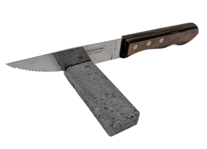 The Black Rock Grill Lava Rock Knife Rest – Limited Edition Table Accessory by Black Rock Grill holds a kitchen knife with a wooden handle, its serrated blade resting securely against the volcanic lava stone.