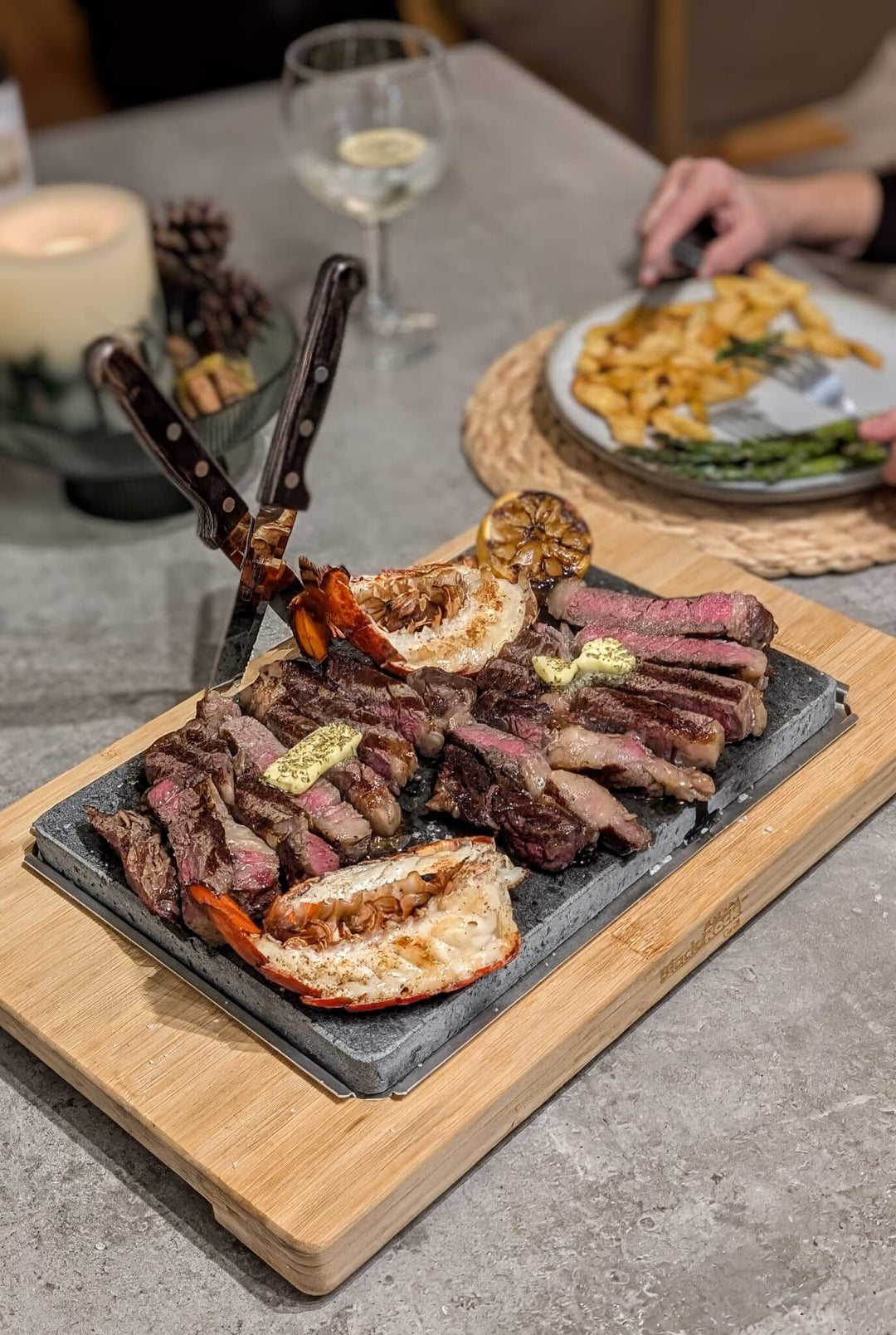 A Black Rock Grill Large Lava Sizzler Finishing Warming Platter holds sliced steak topped with butter, grilled lobster halves, and a charred lemon. Two knives cross the steak as fries and asparagus are served in the background.