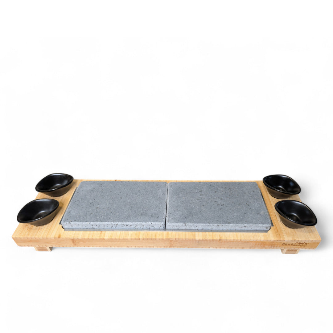 The Sharing Lava Sizzler Finishing Warming Platter by Black Rock Grill features a rectangular bamboo base with two natural lava stone slabs in the center and four small black bowls at each corner, set against a plain white background.