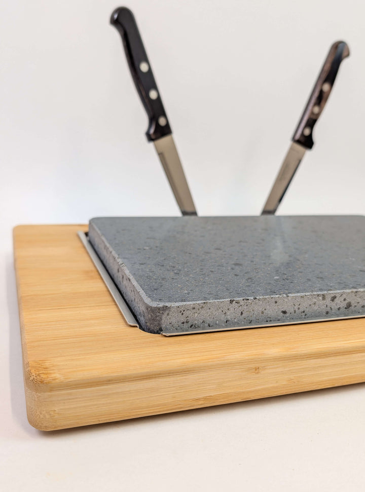 The Black Rock Grill Large Lava Sizzler Finishing Warming Platter features a wooden board with a gray lava stone slab and upright slots for two kitchen knives, making it a stylish, functional serving piece.