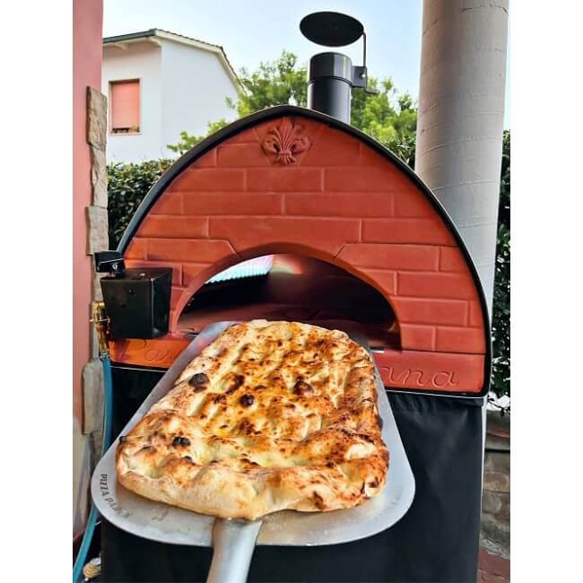 A freshly baked focaccia bread being removed from the Pizza Party Passione dual-fuel outdoor pizza oven on a patio.