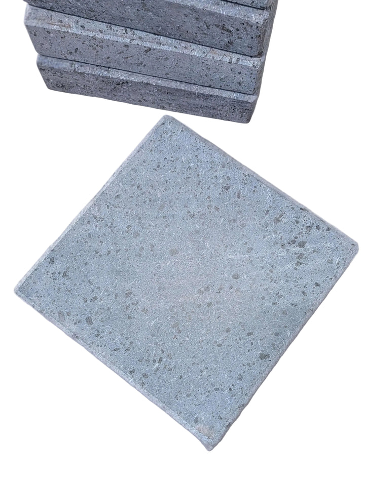 Square gray lava cooking stones with a speckled texture, with one standing in the foreground and a stack of them blurred in the background on a white surface.