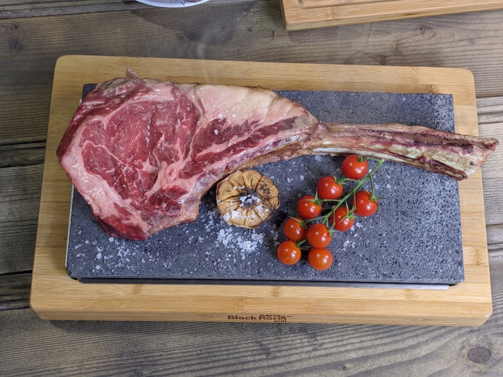 A raw tomahawk steak is being cooked on a lava cooking stone seated on a bamboo board, a sliced garlic bulb, and a cluster of cherry tomatoes, all set on a rustic wooden table.