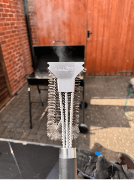 A Black Rock Grill Heavy-Duty BBQ Grill Brush & Scraper is held near the camera, displaying its stainless steel bristles with a smoking grill and brick wall in the background.