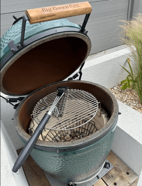 A green ceramic Big Green Egg grill with its lid open features a Black Rock Grill Heavy-Duty BBQ Grill Brush & Scraper (18” stainless steel) resting on the metal grate. The grill is on a patio surrounded by greenery and stones.