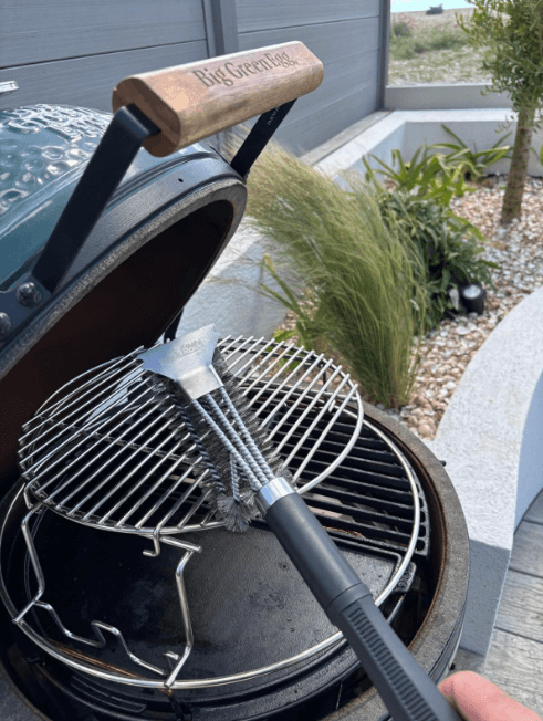 A Black Rock Grill Heavy-Duty BBQ Grill Brush & Scraper, made of 18” stainless steel, cleans the grate of a Big Green Egg barbecue set outdoors near rocks and tall grasses; includes a bonus marinating oil brush.