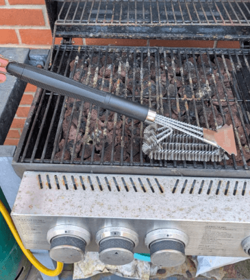The Black Rock Grill Heavy-Duty BBQ Grill Brush & Scraper with stainless steel bristles rests on a gas grills grates, cleaning debris and rust. Control knobs and a yellow hose are visible. Includes a bonus marinating oil brush.