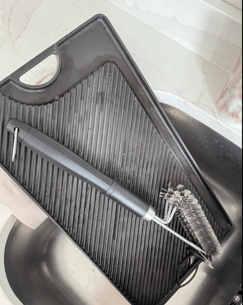A black grill pan sits on a drying rack above the sink, with the Black Rock Grill Heavy-Duty BBQ Grill Brush & Scraper (18 stainless steel) resting diagonally on top. A marble countertop is visible in the background.