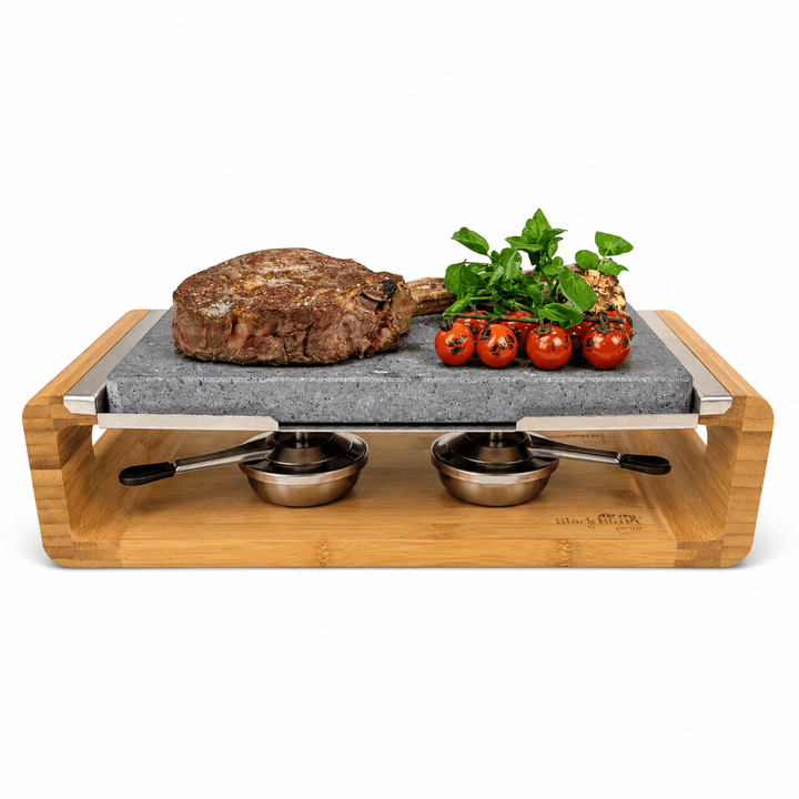 A bamboo-framed black rock grill lava stoner cooking set holding a cooked steak, basil, and cherry tomatoes, with two small metal bowls underneath on a white background.