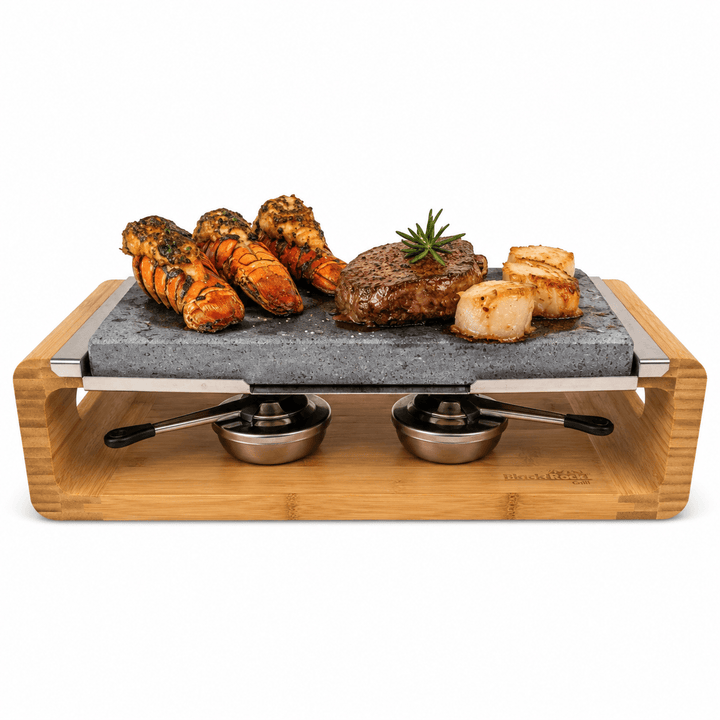 A bamboo-framed tabletop hot lava stone cooking set stove with two stainless steel gel burners to keep the stone hot for longer, photographed on a clean white background., shown with cooked lobster tails, a steak, scallops, and a sprig of rosemary on a white background.