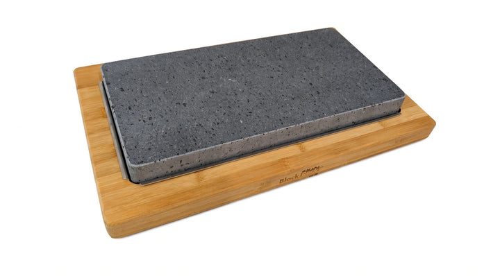 A rectangular dark gray lava cookinf stone resting on a light natural bamboo block, photographed on a clean white background.