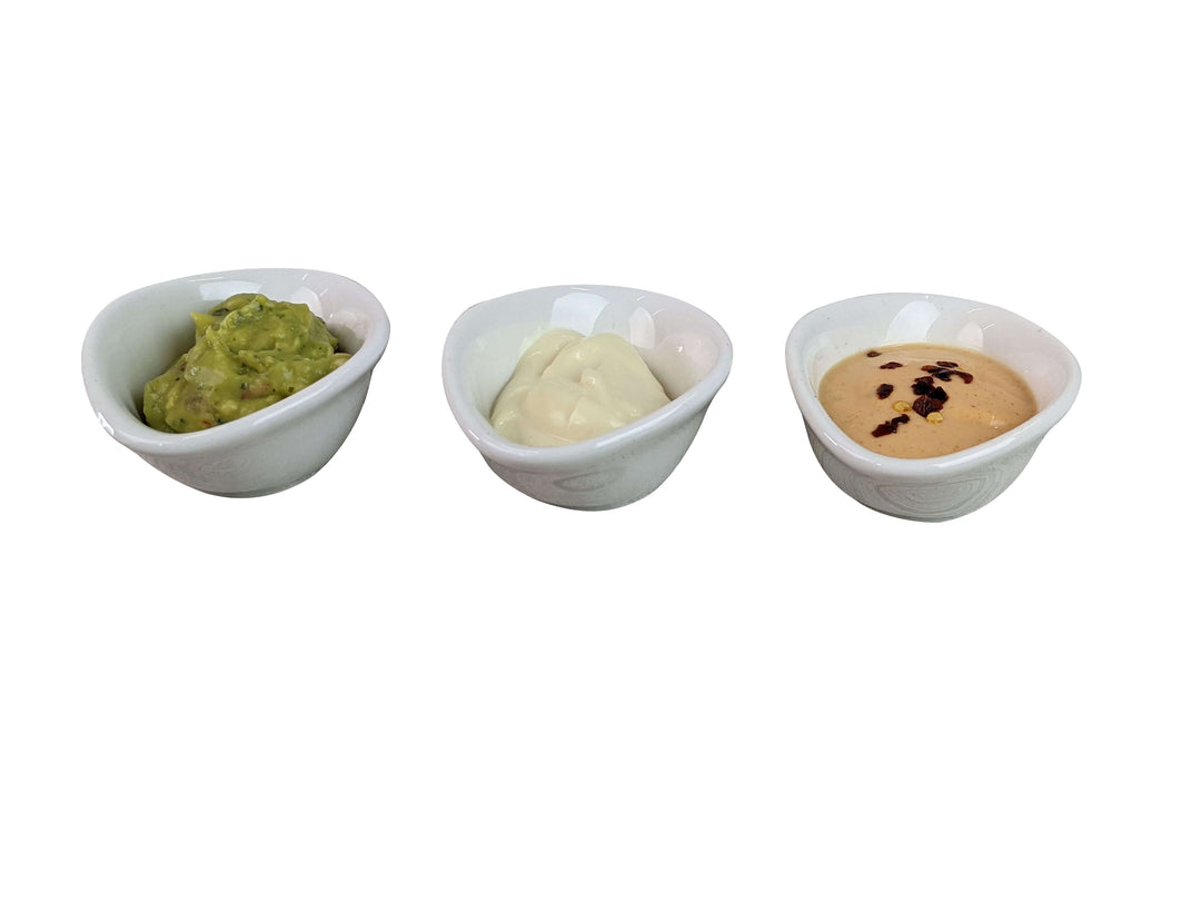 Three small white ceramic oval dipping pots containing green guacamole, white sour cream, and a tan peanut sauce with black chili, arranged in a row on a white background.