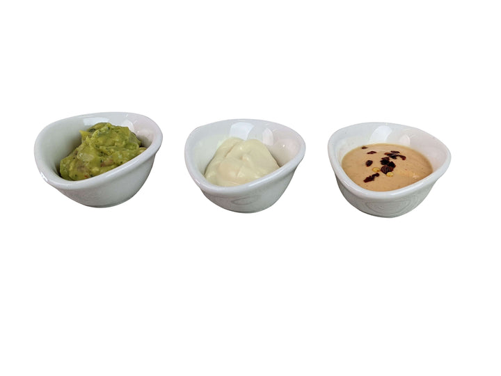 Three small white ceramic oval dipping pots containing green guacamole, white sour cream, and a tan peanut sauce with black chili, arranged in a row on a white background.