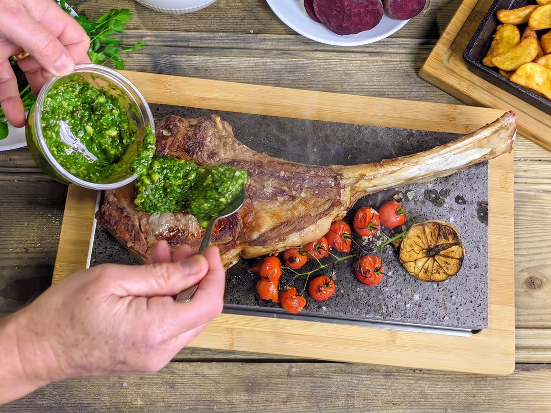 A close-up of a tomahawk steak on a lava cooking stone seated on a bamboo board, with a hand holding a small glass bowl of green herb sauce and spooning it over the meat cherry tomatoes, a garlic slice, and side fries are arranged around on a wooden board over a rustic wood table.