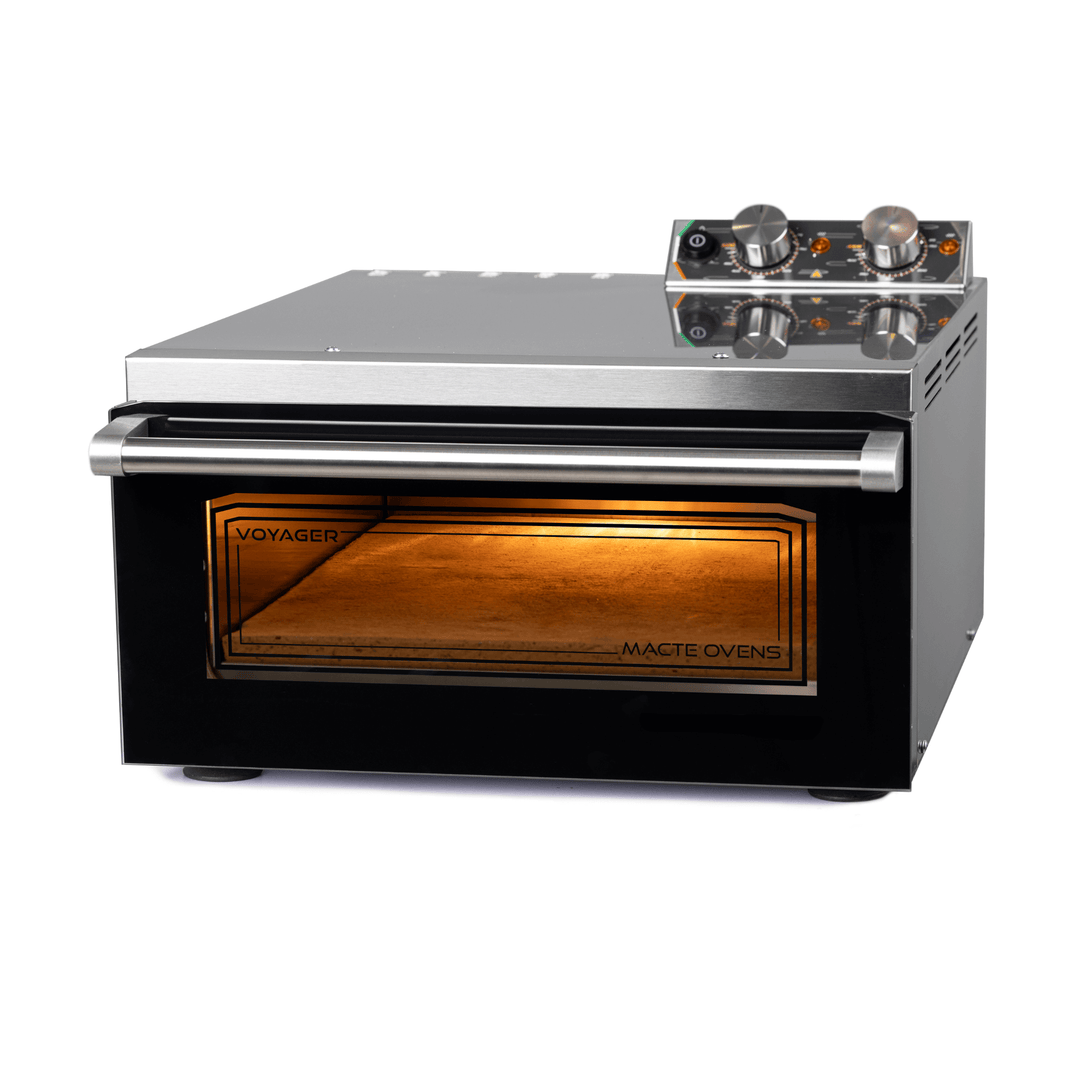 The Macte Voyager Twin Electric Pizza Oven is an AVPN Certified Italian countertop oven with a stainless steel body, glass front door featuring "VOYAGER" and "MACTE OVENS," two control knobs, and an illuminated interior.