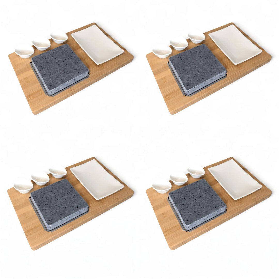 Four identical sets of a bamboo boards with a lava cooking stone, a white rectangular side plate, and three small white dipping pots, arranged in a 2x2 grid on a white background.