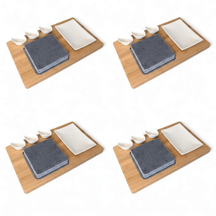 Four identical sets of a bamboo boards with a lava cooking stone, a white rectangular side plate, and three small white dipping pots, arranged in a 2x2 grid on a white background.
