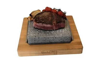 The Black Rock Grill ROXY45 set features a hot stone on a wooden board, allowing you to cook steak with grilled tomatoes and onions at your table for a unique live cooking experience.