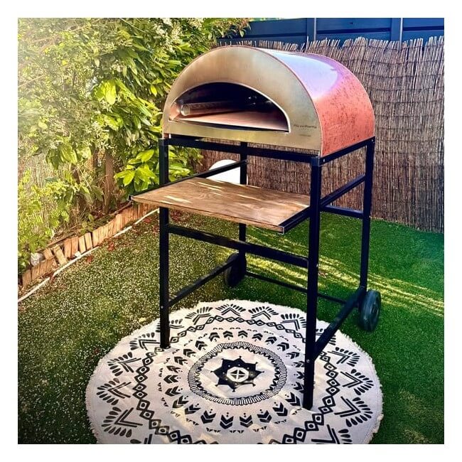 A Pizza Party Black Pizza Party Outdoor Pizza Oven Stand, designed for 70x70, Emozione & Ispirazione ovens, holds a portable pizza oven with convenient wheels on a patterned round rug amidst artificial grass and leafy garden plants, providing sturdy support for outdoor cooking and easy mobility.