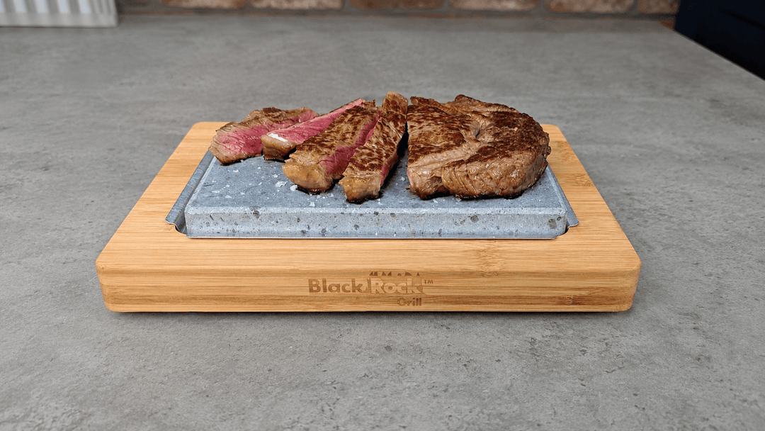 A partially cooked and sliced steak sizzling on a hot warming lava stone set. The warming hot rock sits on an underplate and bamboo serving board on a grey rustic kitchen surface.