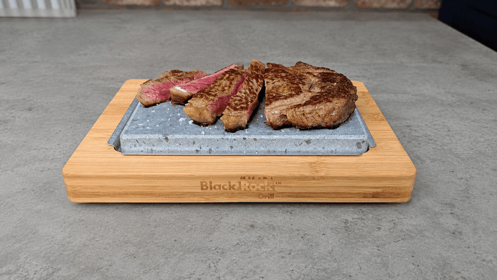 A partially cooked and sliced steak sizzling on a hot warming lava stone set. The warming hot rock sits on an underplate and bamboo serving board on a grey rustic kitchen surface.