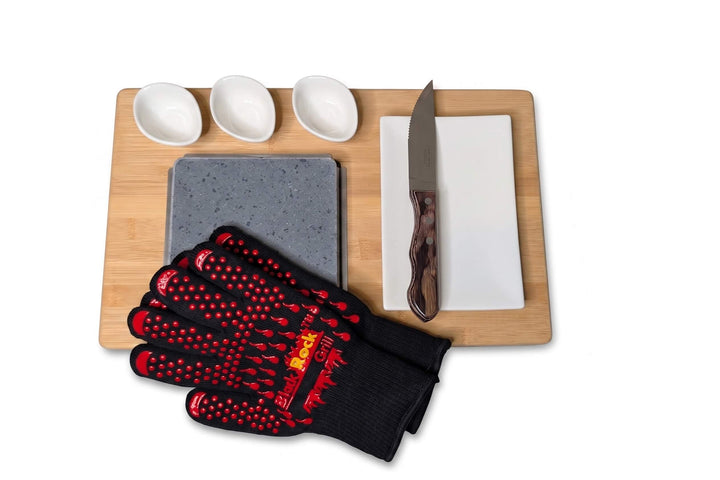 A bamboo serving board set on a white background, featuring a square lava cooking stone with a pair of black rock grill heat resistant gloves placed over one corner, a rectangular white side plate with a steak knife on it and three small white dipping pots.