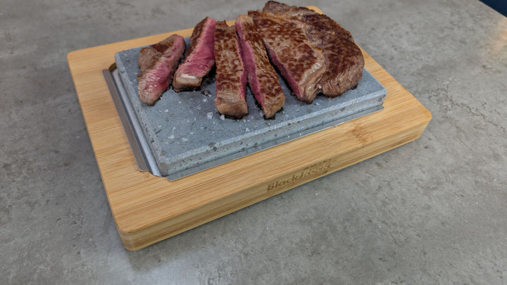 Sizzling steak finishing cooking on an individual hot lava stone warming set, featuring a bamboo board and underplate, placed on a rustic grey sideboard.