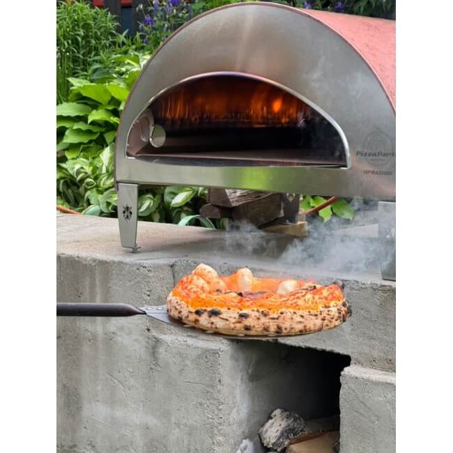 A freshly cooked pizza resting on a peel in front of a Pizza Party Ispirazione outdoor oven in a garden setting.
