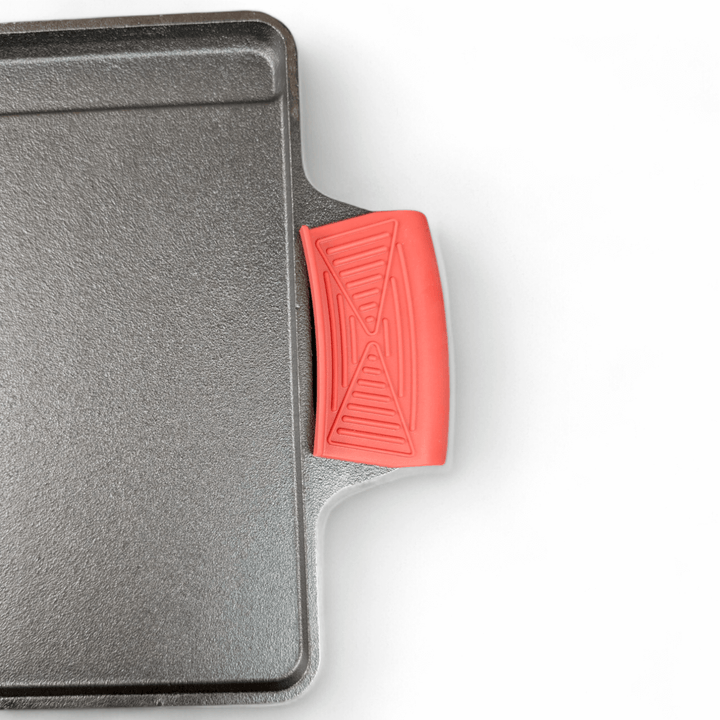 A close-up of a black cast iron griddle features its empty cooking surface with a Black Rock Grill silicone heat-resistant handle cover. The food-grade silicone ensures safe usage, and the white background accentuates the rectangular shape and textured finish of the griddle.